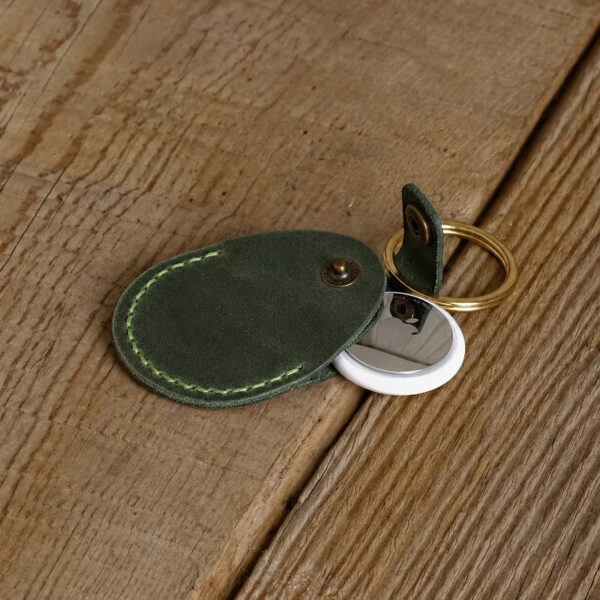 Green full-grain leather AirTag holder with an AirTag inside and a brass key ring, on a wooden table