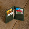 Green full-grain leather six-pocket card wallet opened on a wooden table, filled with bank cards and folded notes