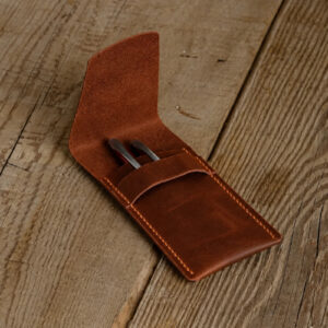 Brown full-grain leather pen case opened on a wooden table with two pens in the pocket