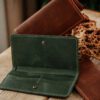 Green full-grain leather long travel wallet opened to show its card slots, brown wallets behind it on a wooden table