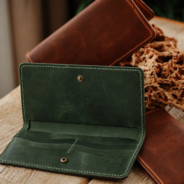 Green full-grain leather long travel wallet opened to show its card slots, brown wallets behind it on a wooden table