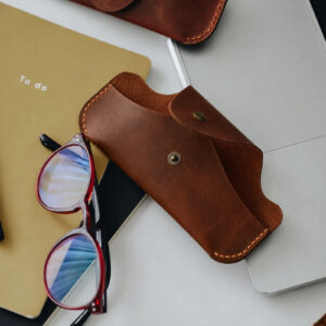 Brown full-grain leather glasses case with a snap flap beside a pair of red glasses and a notebook